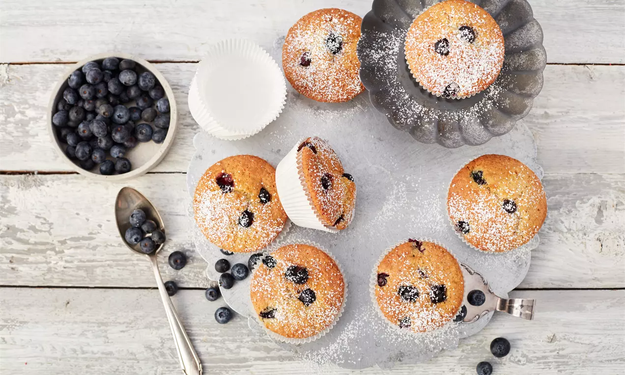 Gyldenbrune blåbærmuffins med dryss av melis på rustikk metallform og lyst trefat, omgitt av friske blåbær, skje og hvite papirformer