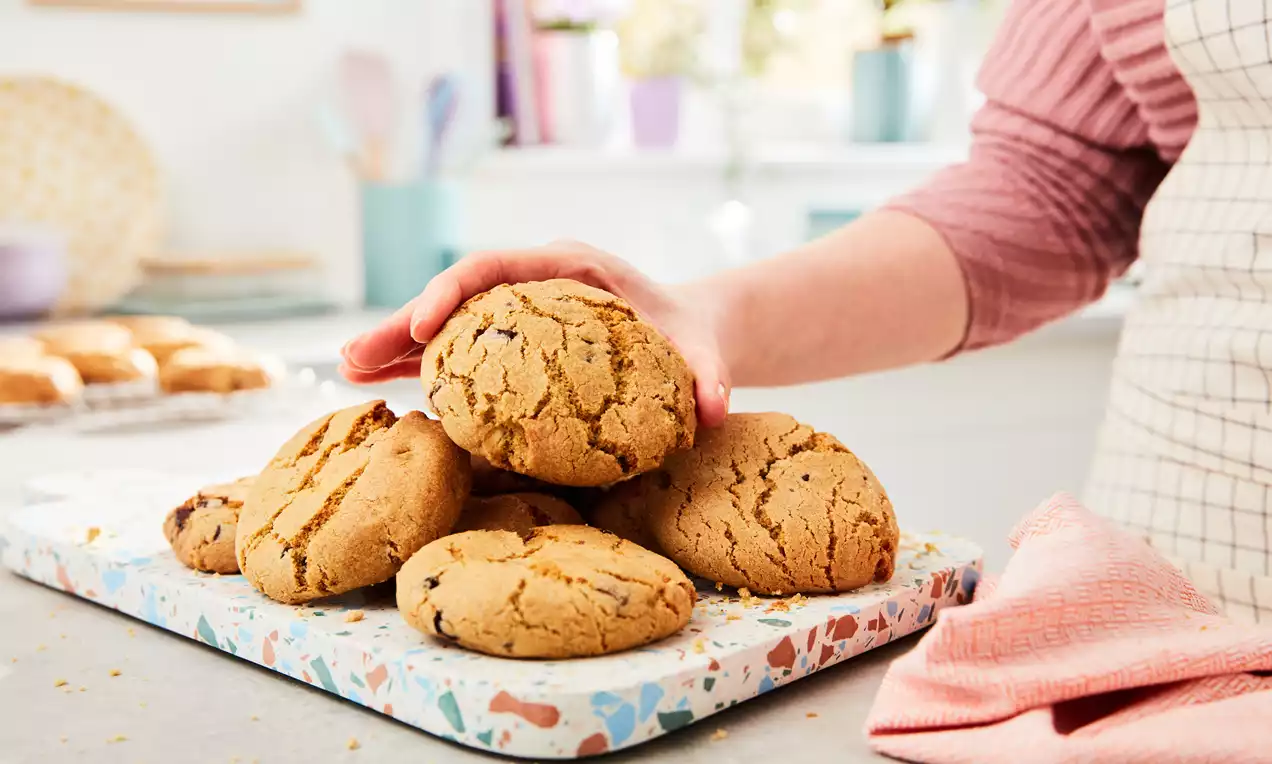 Gyldenbrune cookies med sprukket overflate ligger i en haug på et fargerikt fat, med en hånd som støtter dem og tekstil ved siden av