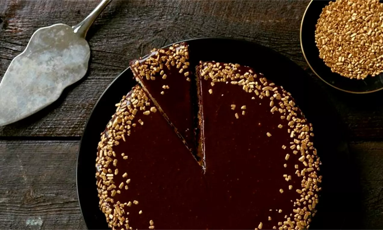 Konfektkake med mørk glasur og gullfarget strøssel langs kanten på et sort fat ved siden av en kakespade og en skål med strøssel