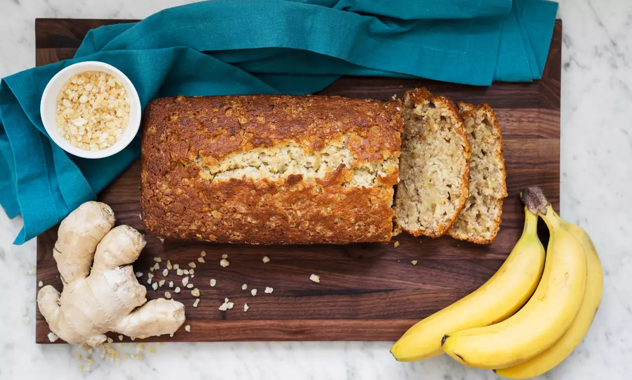 Gyldenbrunt bananbrød med sprø skorpe og saftige skiver på mørkt trefat, omgitt av bananer, ingefær og kandisert ingefær i en hvit skål