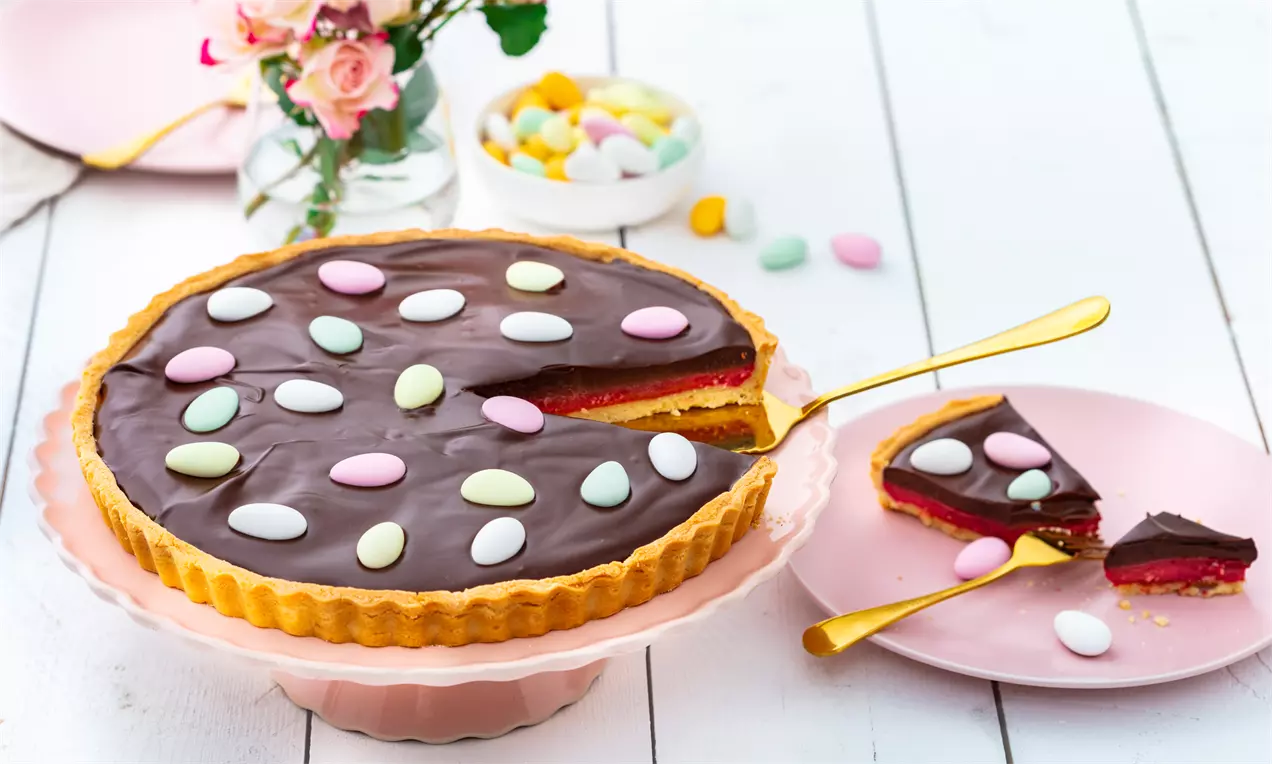 Rund påsketærte med blank mørk chokoladeganache og hindbærcurd, pyntet med lyse påskeegg på rosa kagefad ved siden af pink tallerken