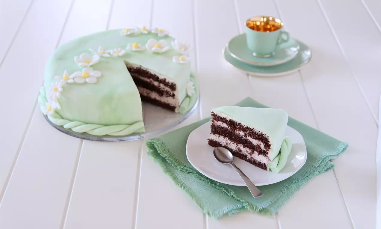 Festkake med lysegrønn fondant dekorert med hvite og gule blomster, en kakeskive på hvit tallerken ved siden av kopp på lysegrønn serviett