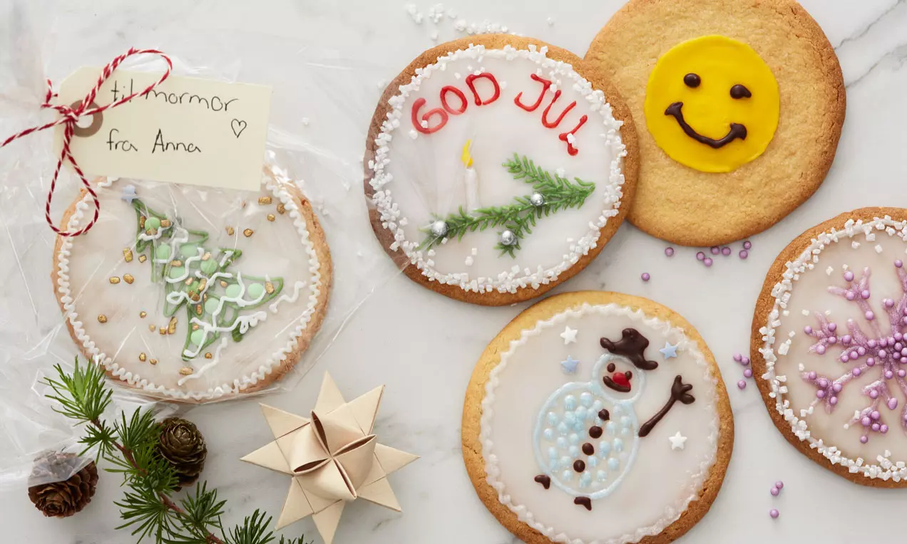 Store, runde småkaker med hvit glasur dekorert med julemotiv som tre, snømann og tekst, plassert på hvit marmor med granbar og stjernefletting