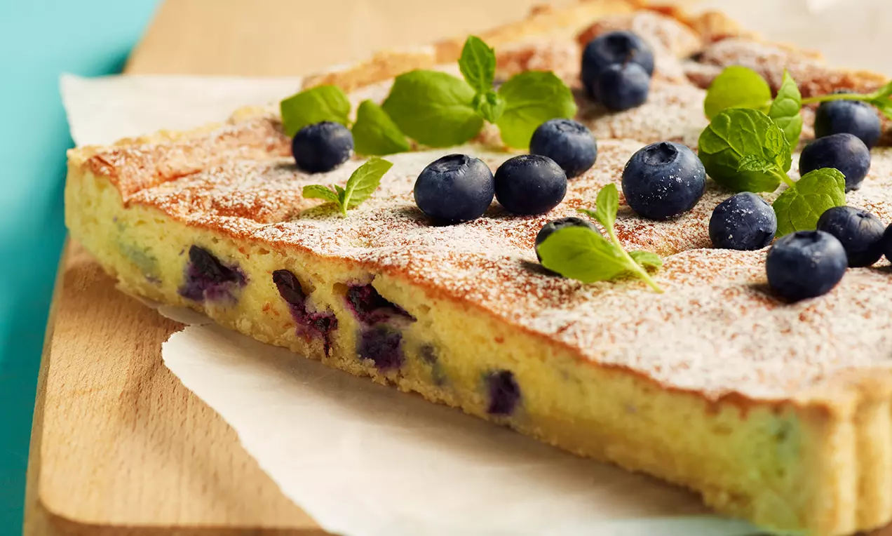 Firkantet blåbærtærte med gyllenbrun kant og lys mandelfyll, toppet med blåbær, grønn mynte og melisdryss på bakepapir