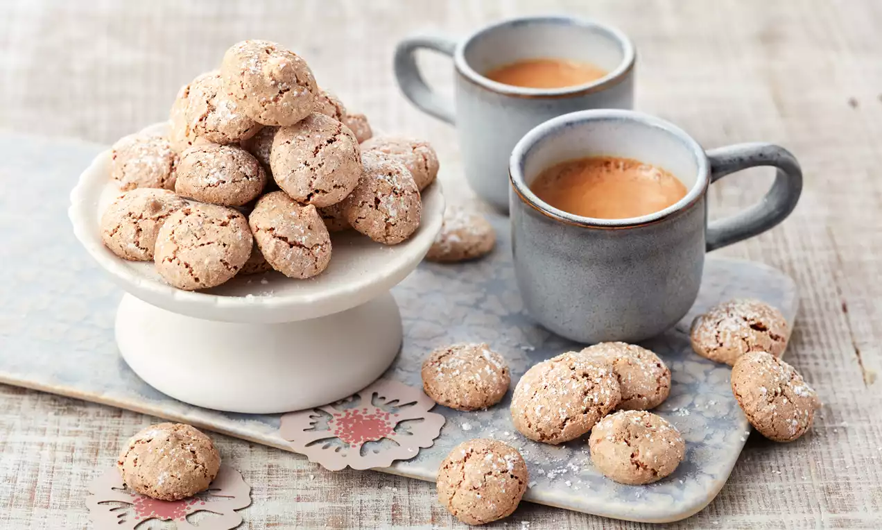 Små, lysbrune amarettini med sprø overflate og dryss av melis, stablet på et hvitt kakefat ved siden av grå kopper med kaffe
