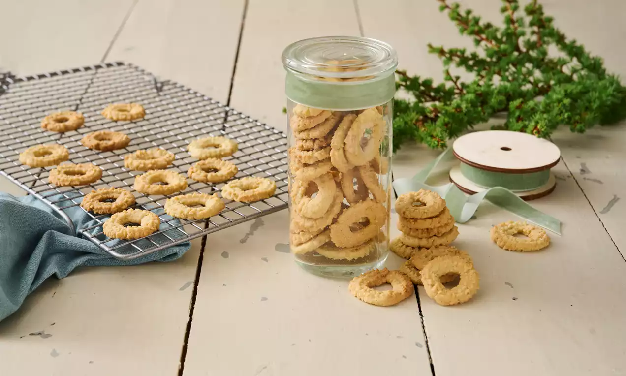 Veganske vaniljekranser gyllenbrune med sprø tekstur formet som runde kranser på rist og i glass omgitt av bånd og grønn einerkvist