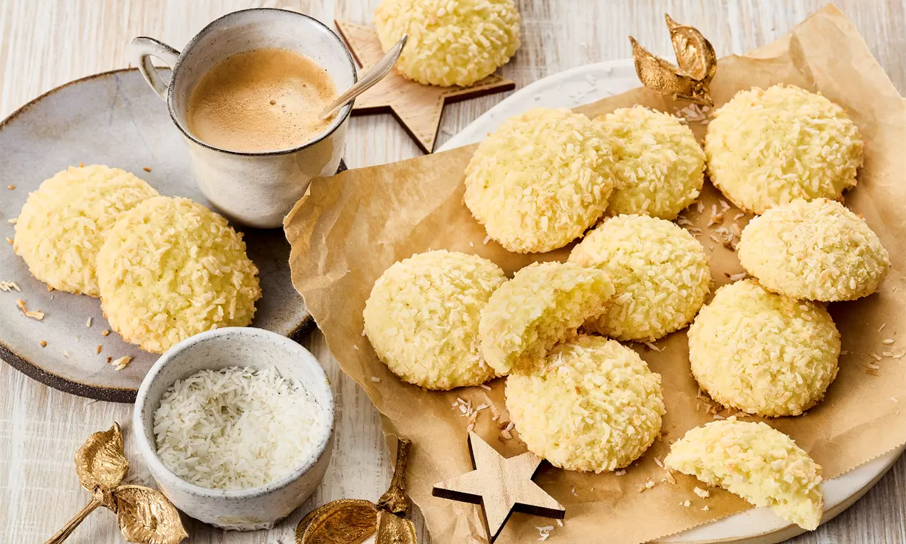 Runde, gylne kokos-cookies med crispy overflate ligger spredt på brunt bakepapir med kokosdryss, kaffekopp og dekorative elementer rundt