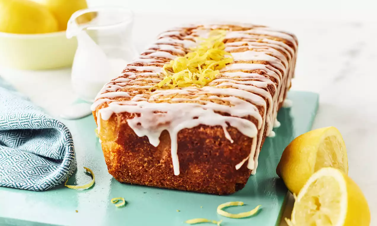 Vegan sitronkake i avlang form med hvit glasur og sitronskall på toppen, plassert på turkis brett med halve sitroner og blått klede