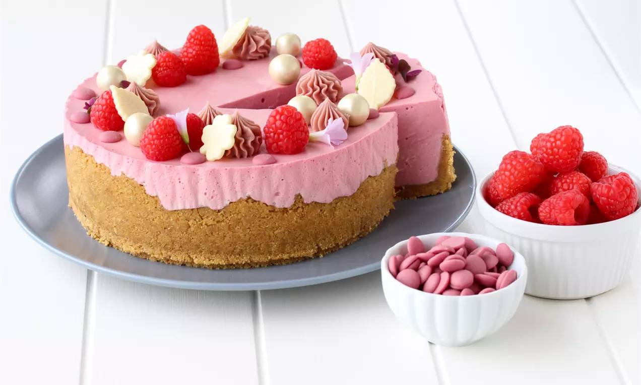 Bringebærostekake med kremet rosa fyll, gyllen kjeksbunn, dekor av bringebær, ruby-sjokolade og kremtopper på et gråblått fat