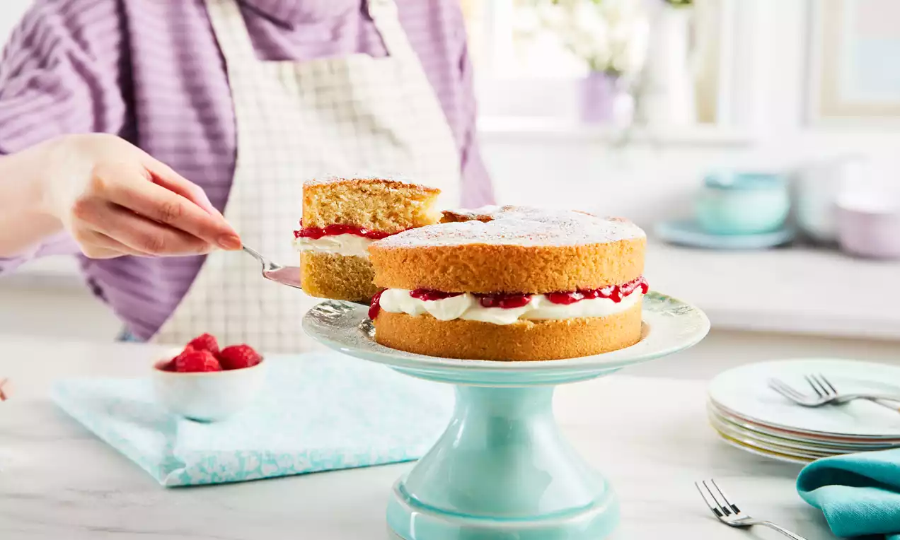 Victoria Sponge med lysebrune lag fylt med krem og røde bær, drysset med melis på lyseblå kakestativ ved siden av skål med bringebær