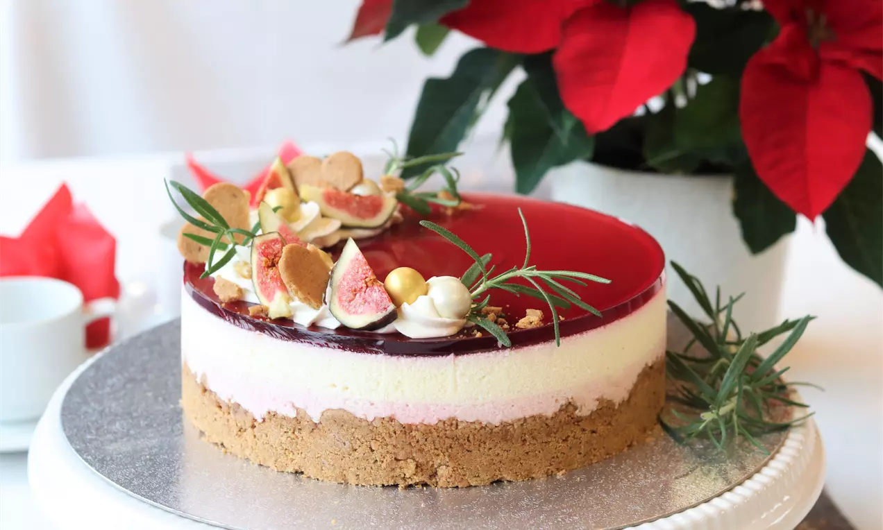 Juleostekake med brun kjeksbunn, lag av hvit og rosa ostekrem, rød gløgggele og pynt av fikener, rosmarin og gullkuler på et kakefat