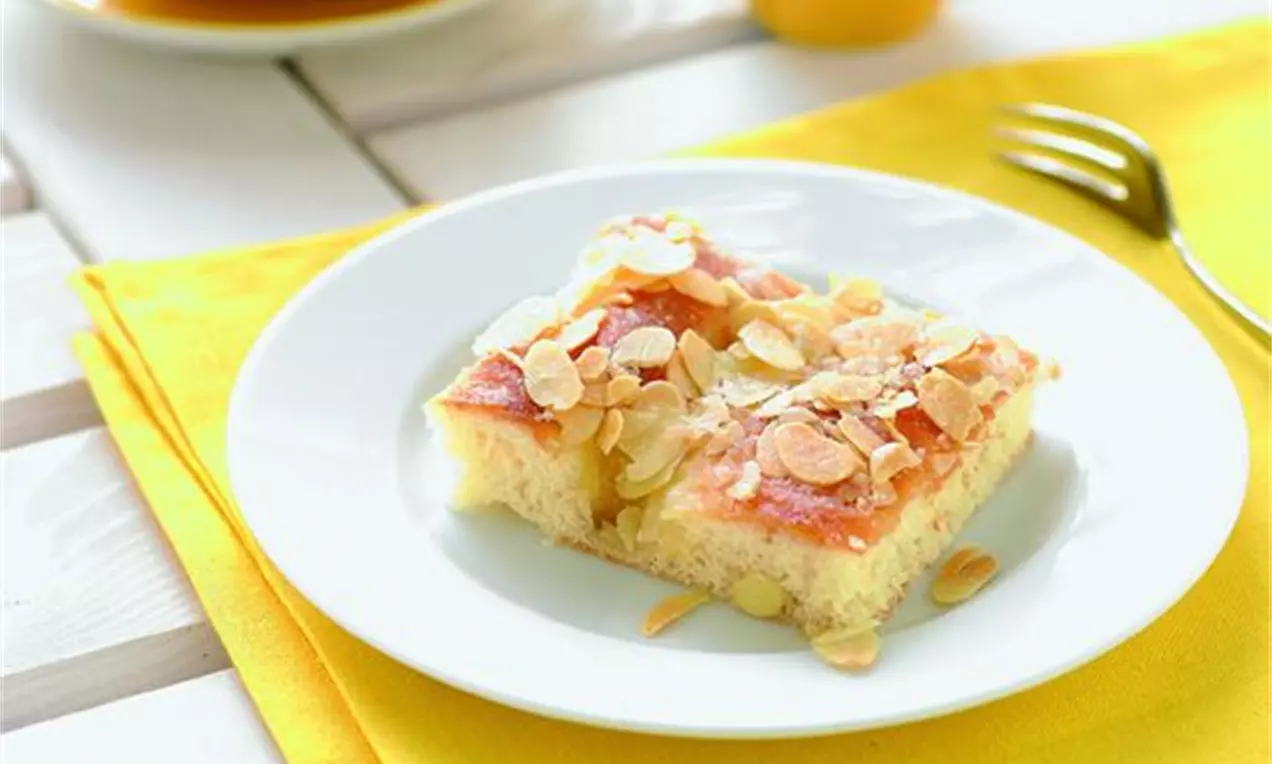 Firkantet smørkake med gyllen overflate og sprø mandelflak på en hvit tallerken på en gul duk med gaffel og kopp i bakgrunnen