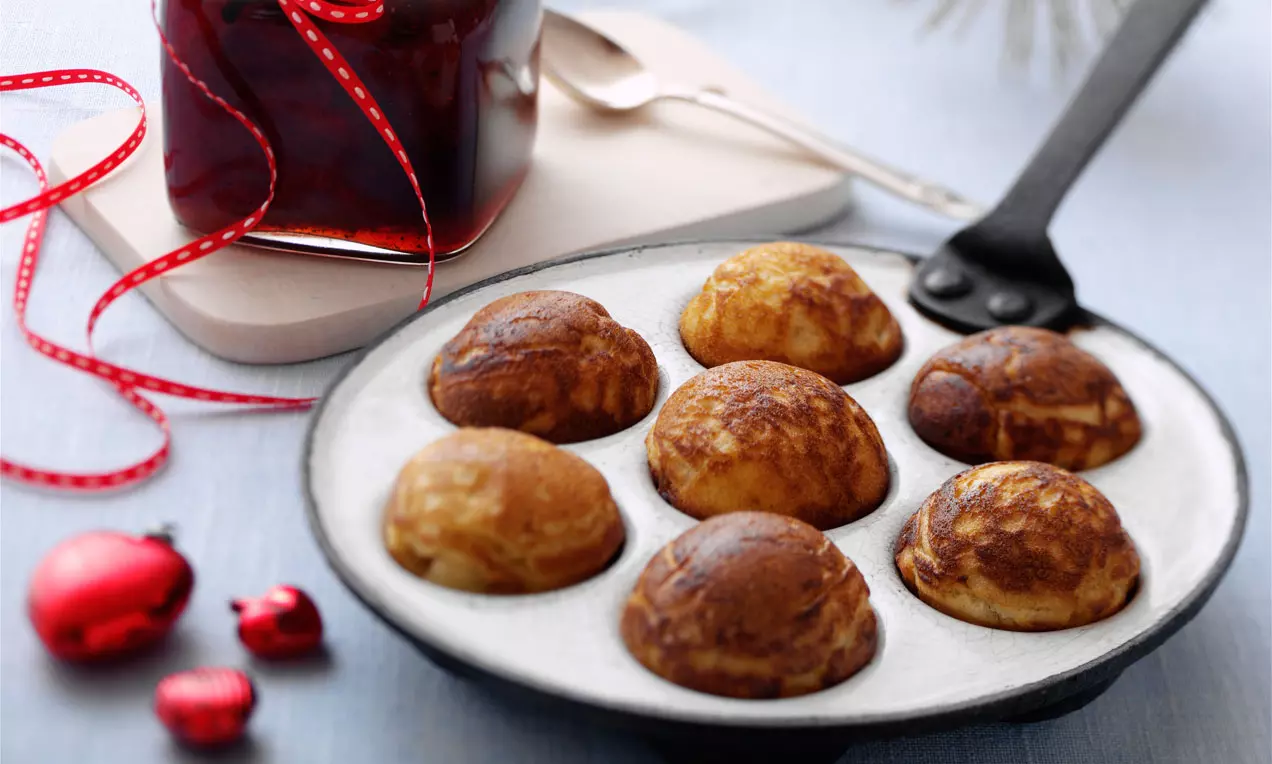 Gyldenbrune runde æbleskiver med sprø overflate i svart støpejernspanne, servert med syltetøy i krukke med rød sløyfe